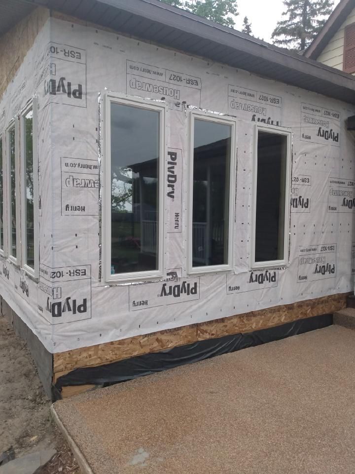 Partially built house wrapped in white housewrap with several windows installed, on a concrete foundation.