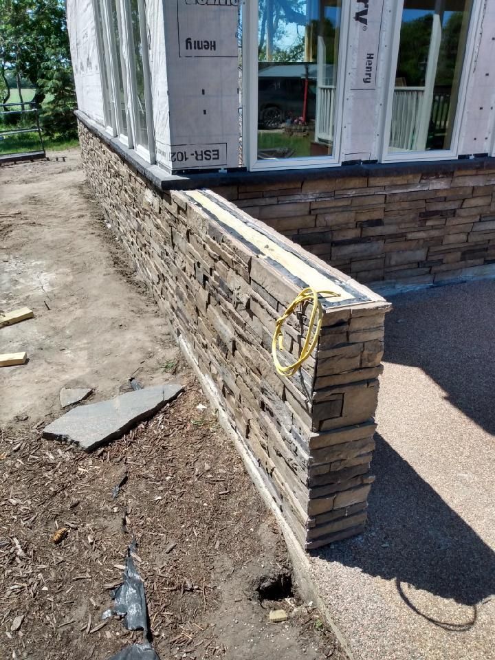 Partially built stone wall beside a house under construction, with exposed framing and dirt ground