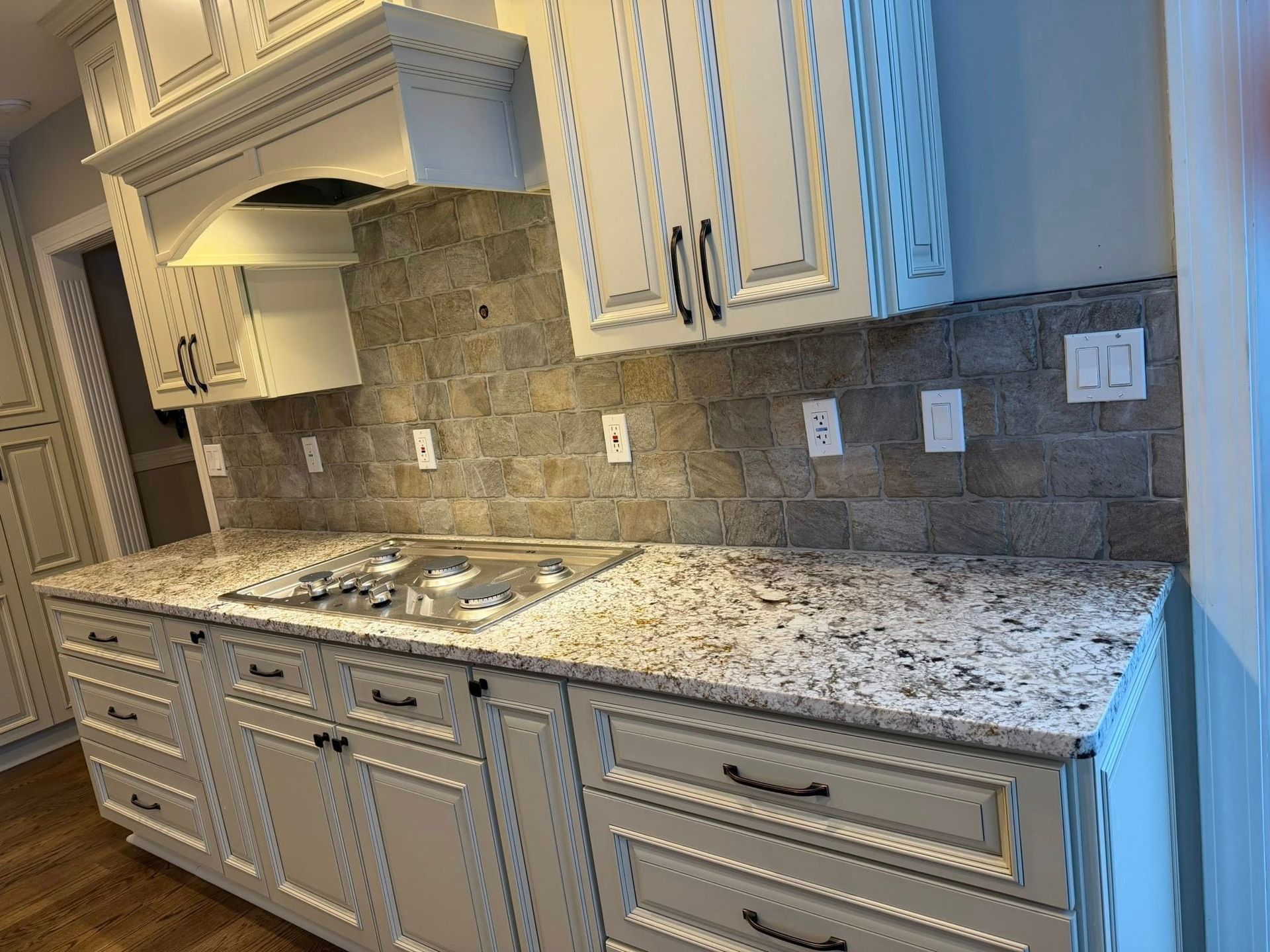 Granite kitchen countertop with white cabinets, tiled backsplash, sink, and stove under a range hood.