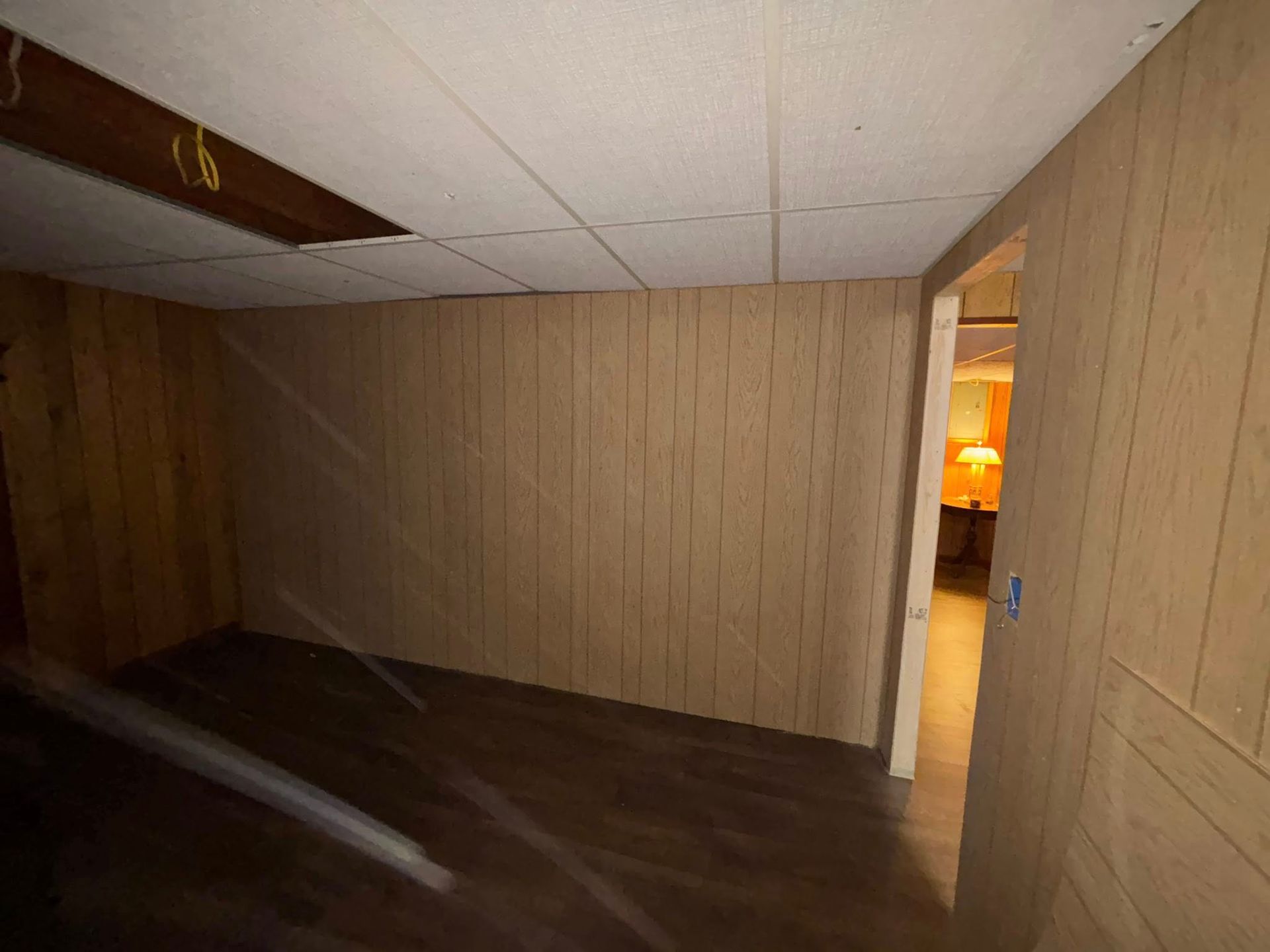 Empty beige-paneled room with damaged ceiling, dark floor, and a lit doorway at the far right