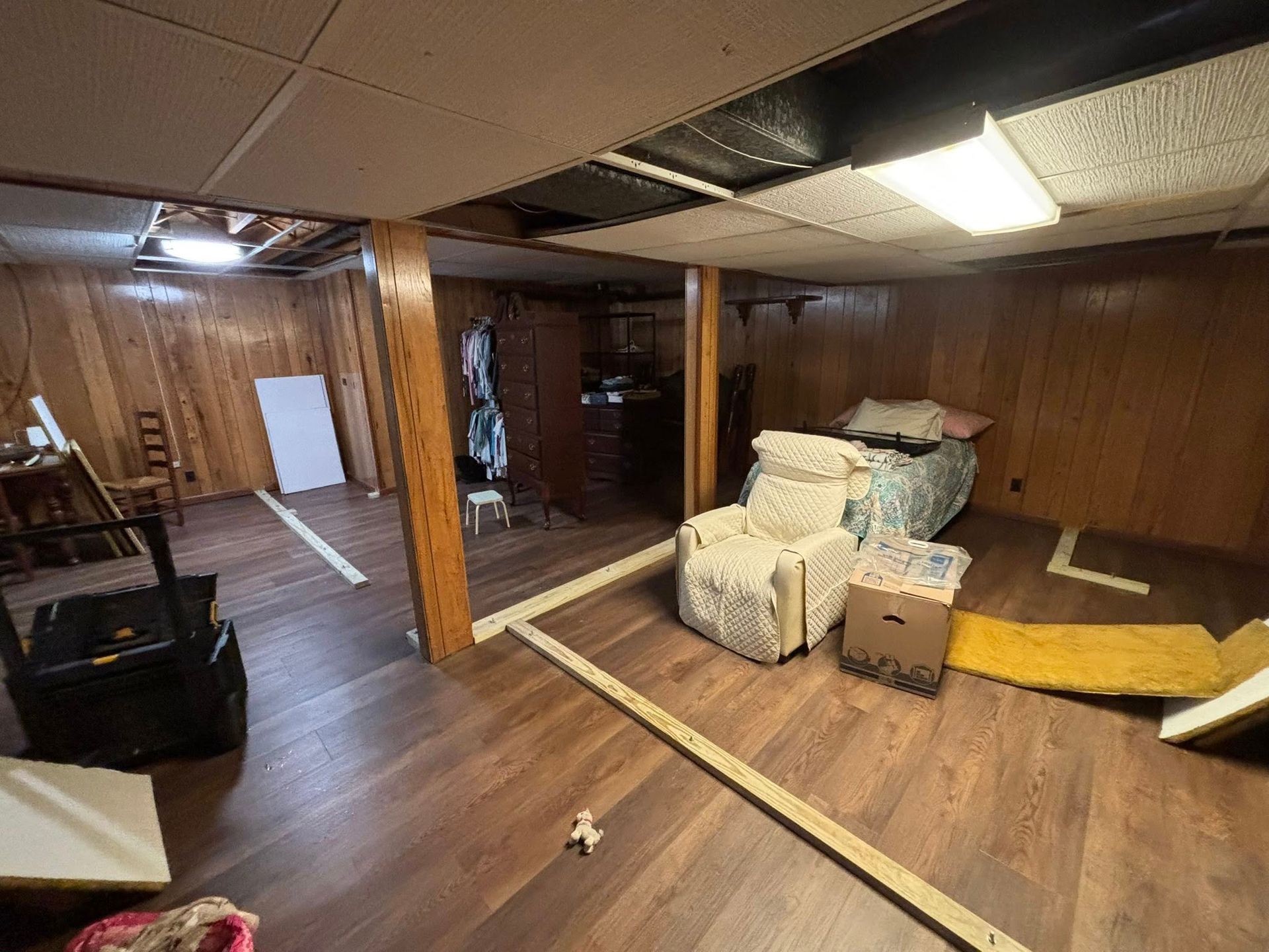 Basement with wood-paneled walls, low ceiling, and scattered furniture and boxes on a wood floor.