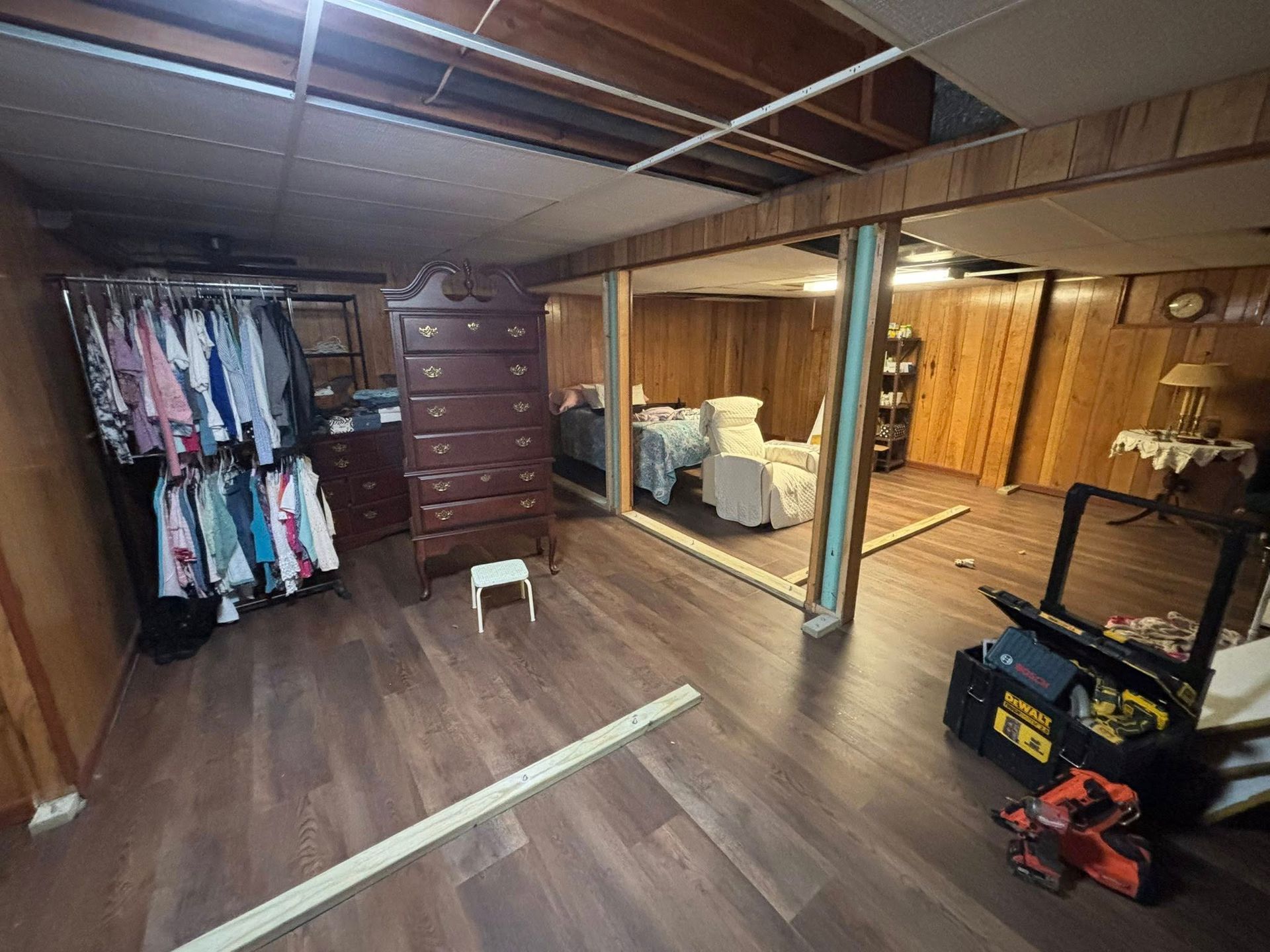 Basement room with wood floor, hanging clothes, dresser, mirror, and tools on the floor.