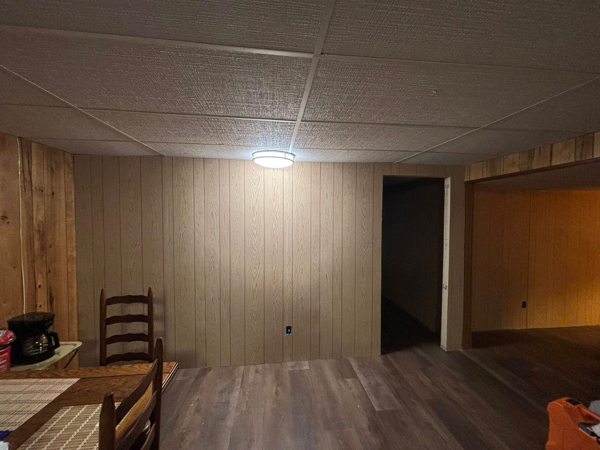 Dim wood-paneled room with a ceiling light, table, chairs, and a dark doorway.