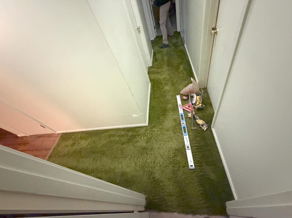 Narrow hallway with green carpet, a measuring tape on the floor, and a hand placing items near a door.