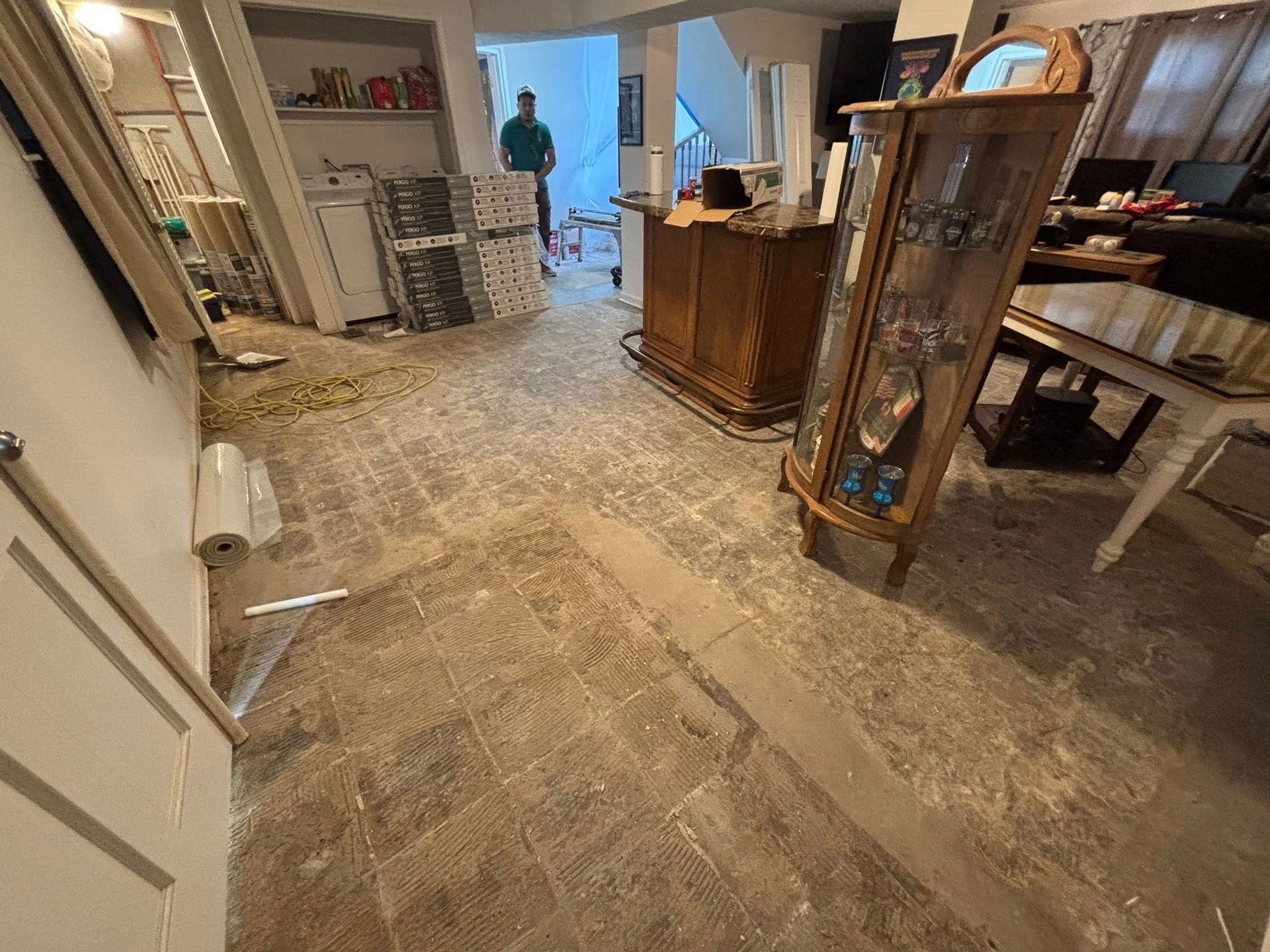 Damaged living room with torn-up floor, scattered debris, and a person in the doorway in the background