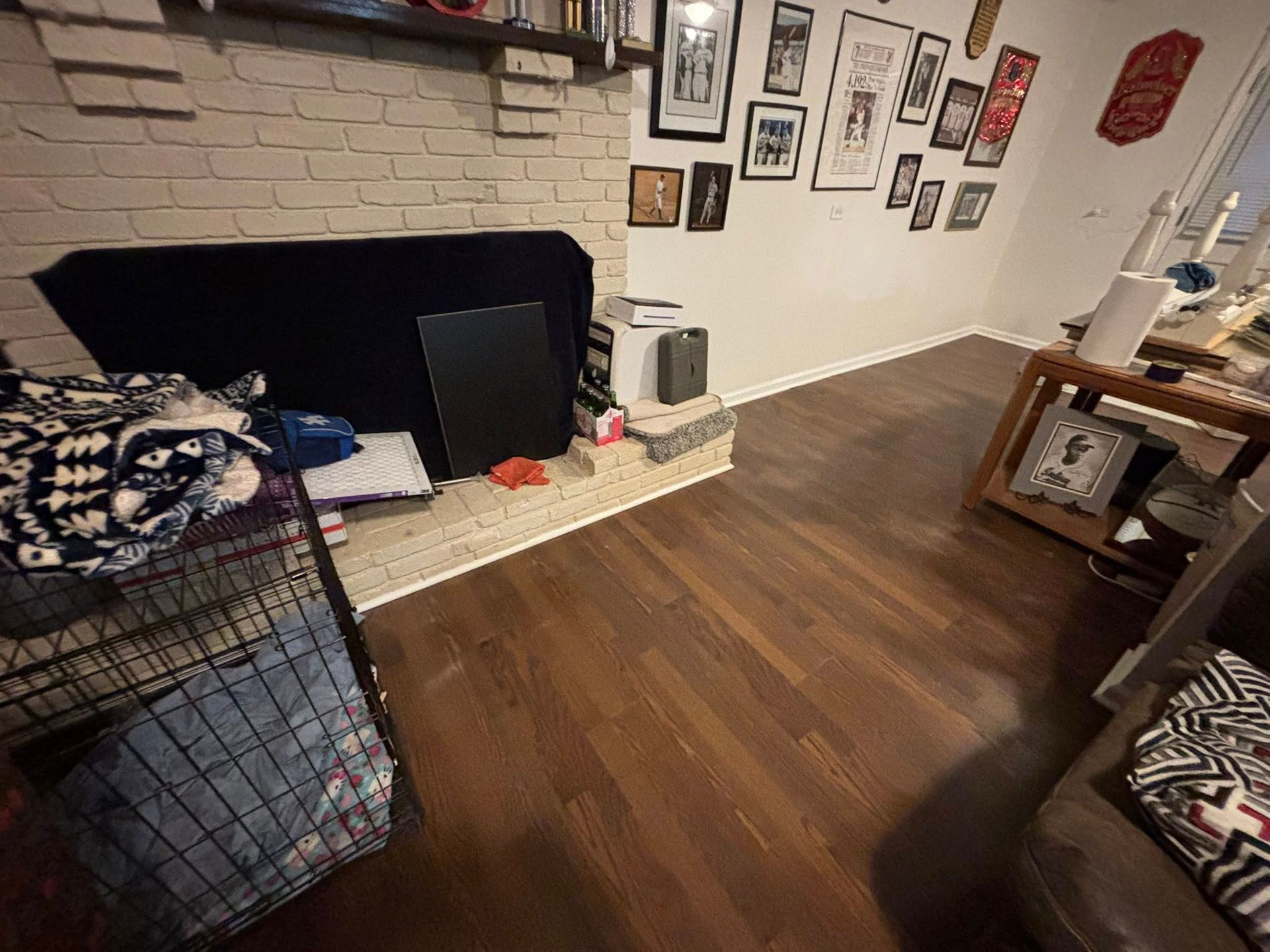 Living room with brick fireplace, hardwood floor, wall art, and pet crates on the left.