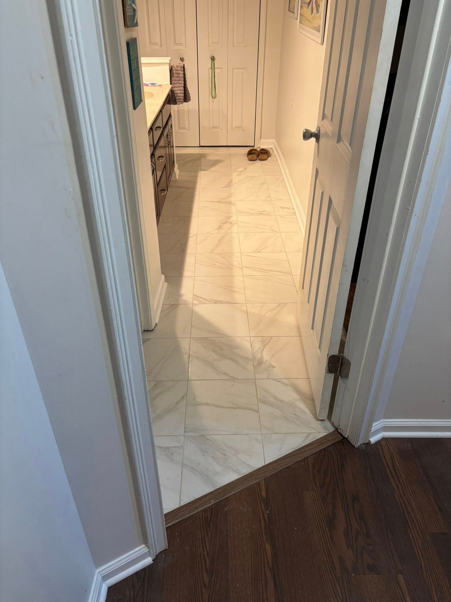 Narrow hallway with white tile floor leading into a bright kitchen, seen from a darker wood floor.
