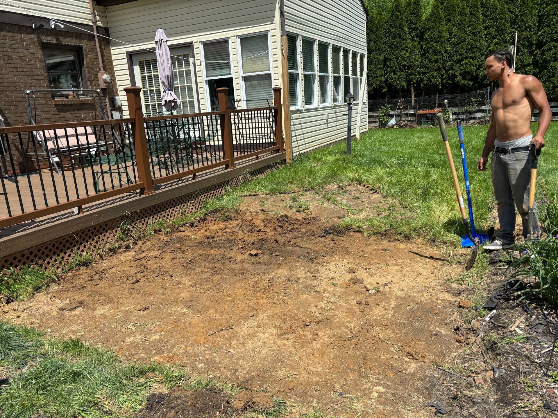Backyard with a dug-up dirt area beside a house deck, with a man holding a shovel on the right.