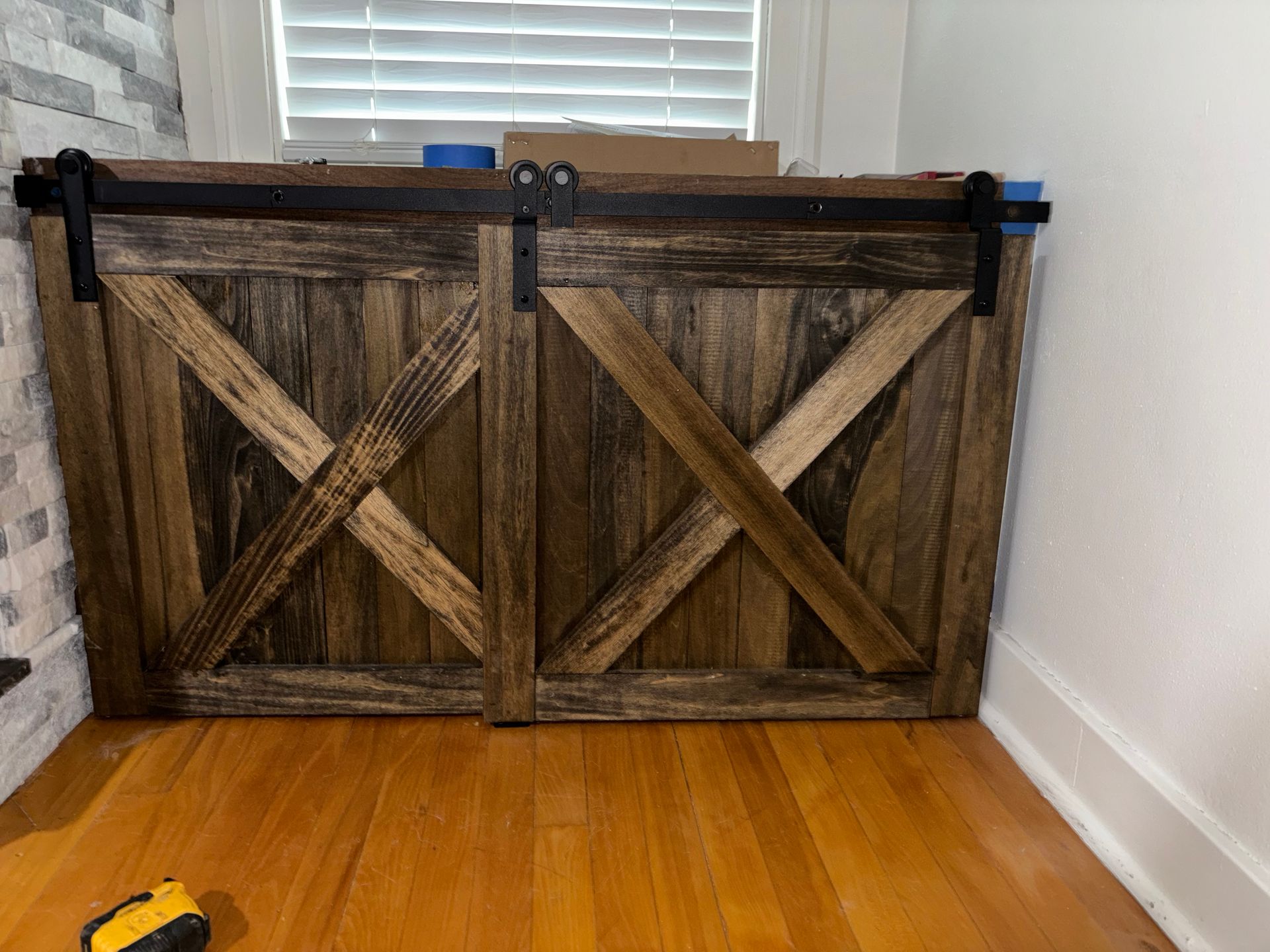 Rustic wooden barn-style cabinet with X doors against a white wall, with a yellow tape measure on the floor.