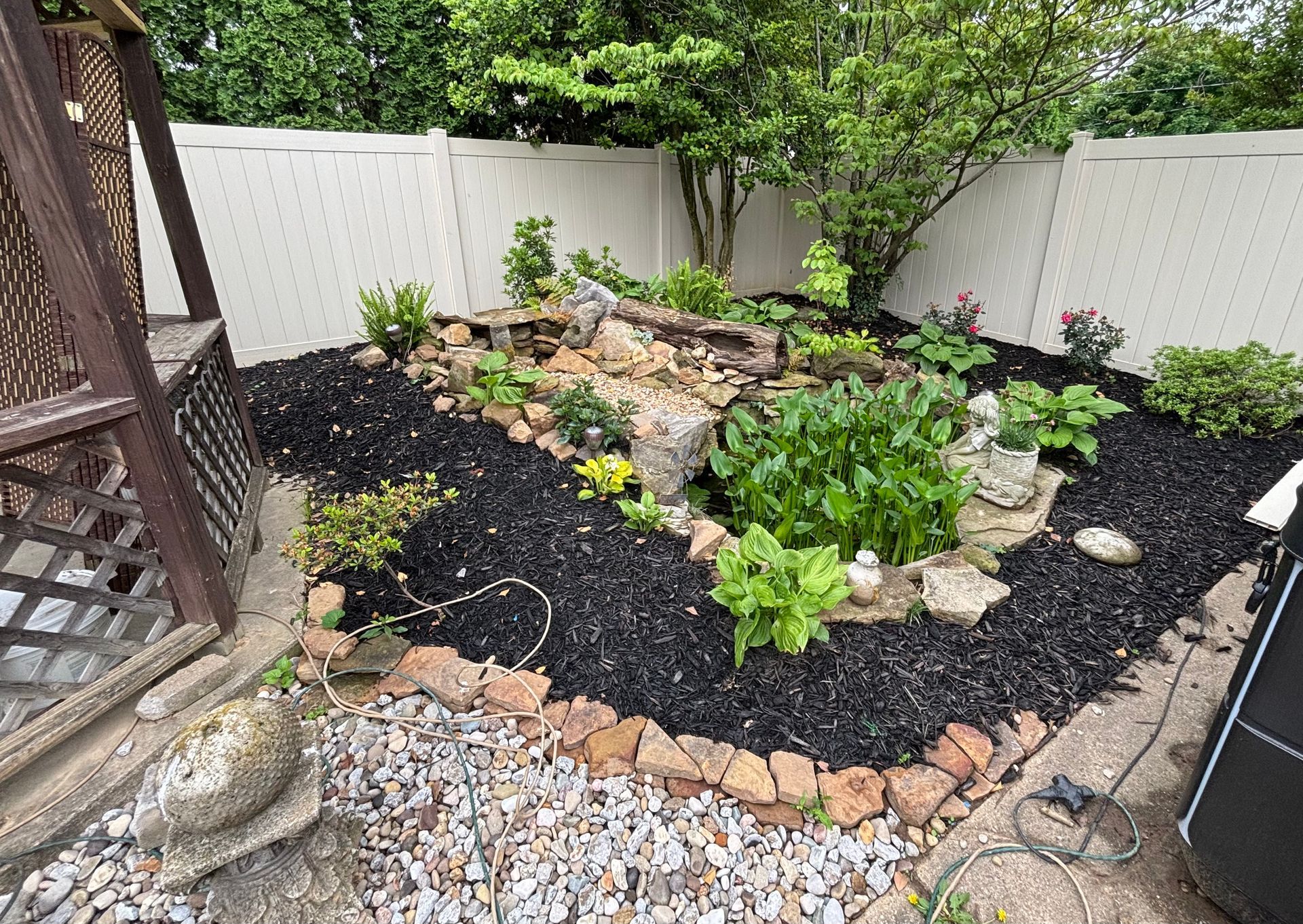 Small backyard rock garden with black mulch, green plants, stones, and a white fence.