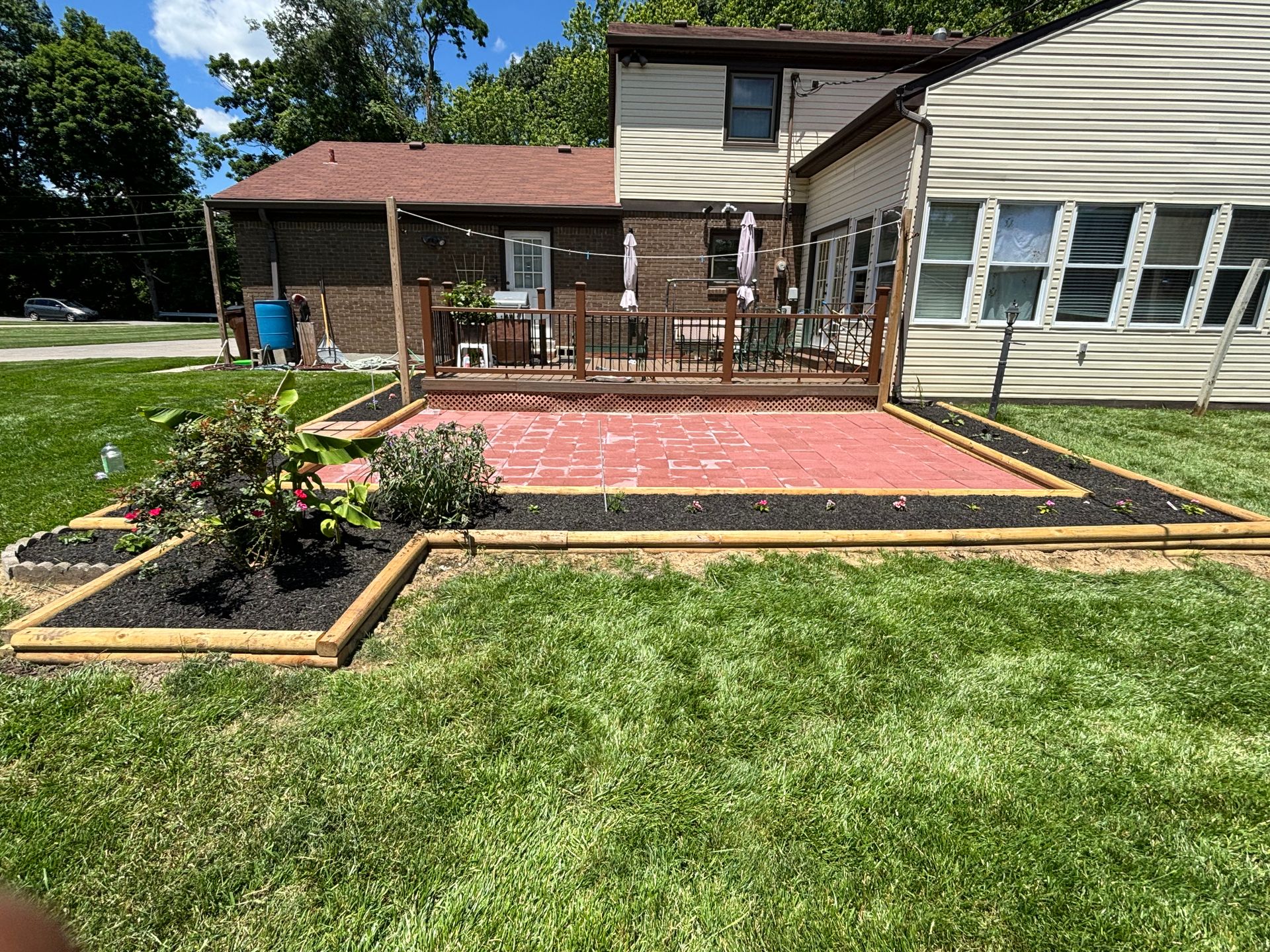 Backyard deck with red brick patio, wooden railing, flower bed, and green lawn beside a house