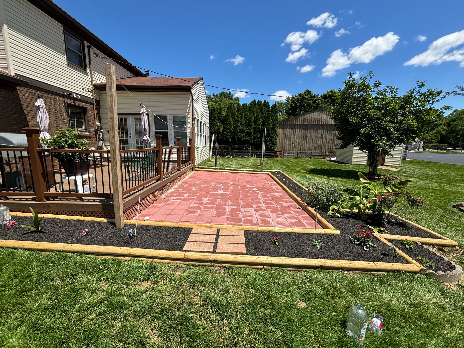 Backyard patio with red pavers, wooden deck railing, and landscaped lawn beside a house