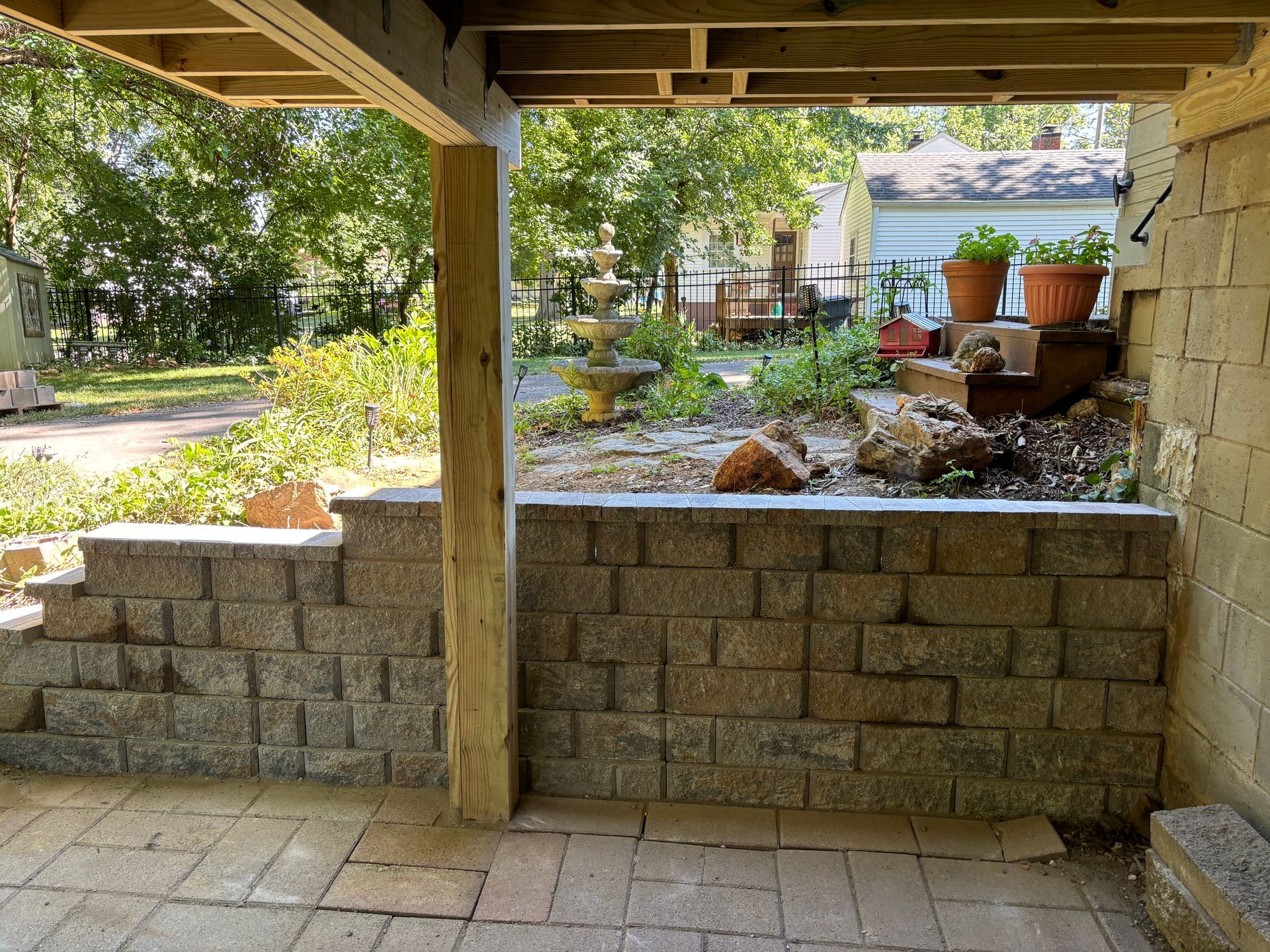Stone patio under a wooden deck, with a retaining wall and a landscaped garden beyond.