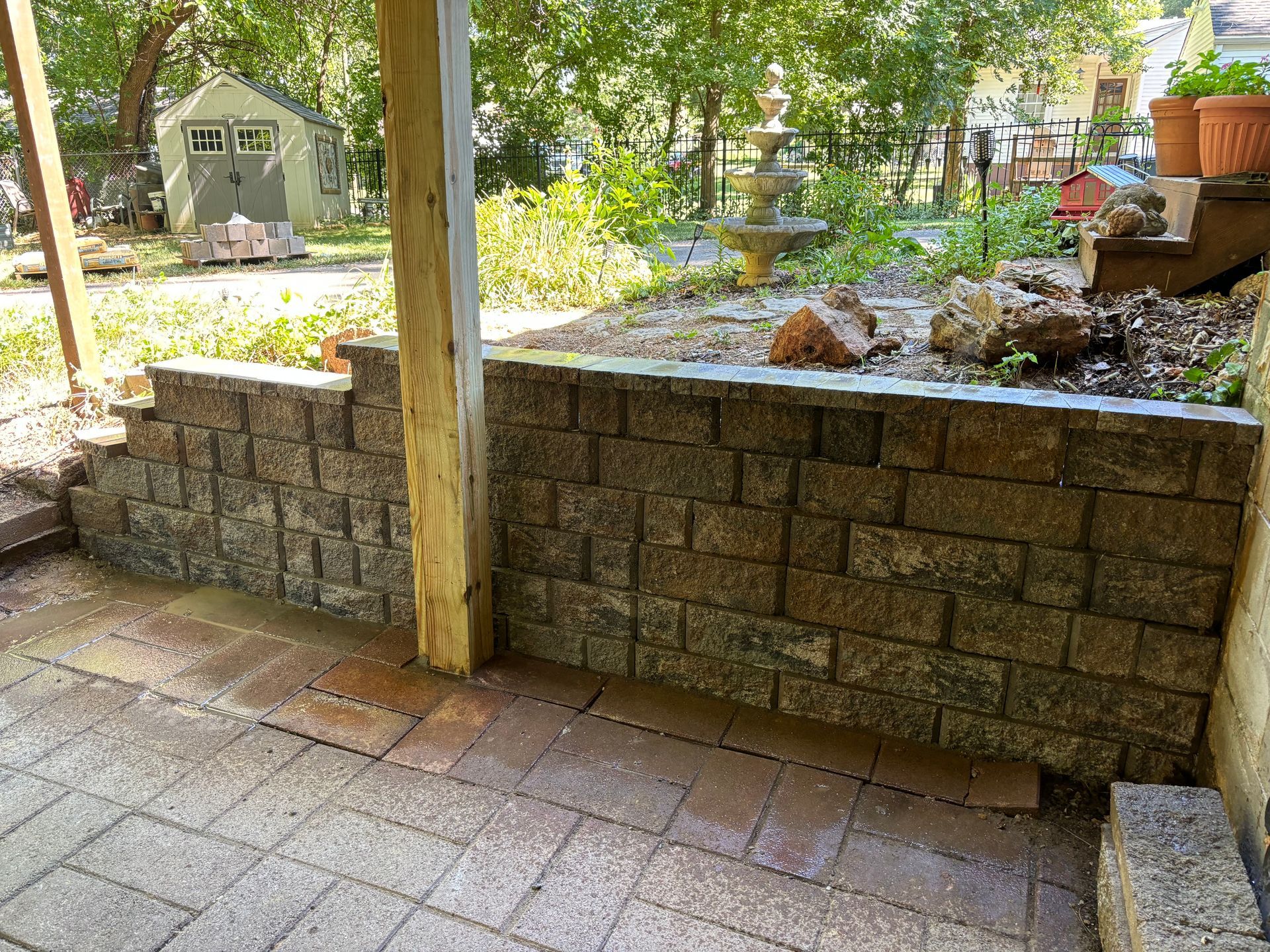Patio with brick pavers, stone retaining wall, wooden post, and garden beds in a sunny backyard