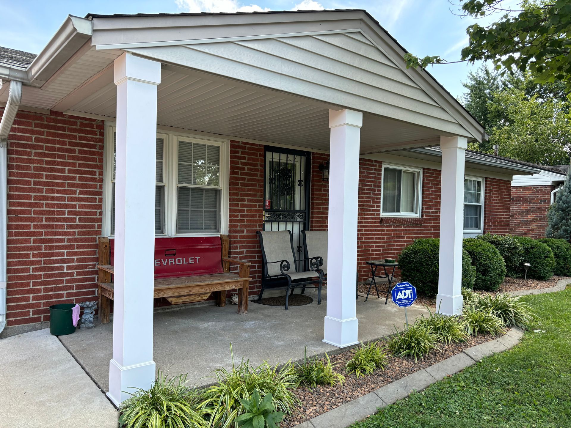 Brick house porch with white columns, red bench, and potted plants