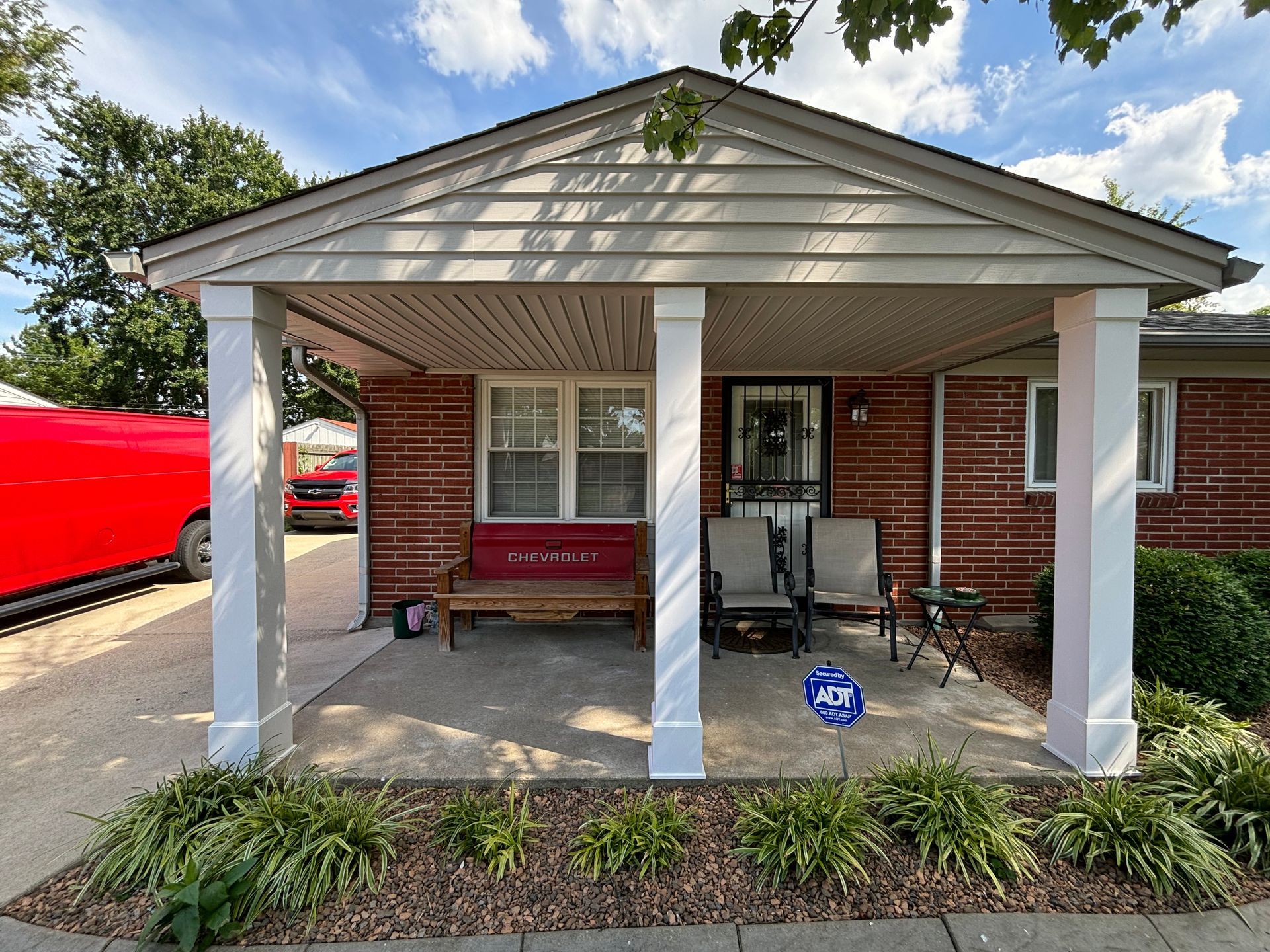 Covered porch with white columns, brick wall, red bench, and a red vehicle parked beside it.
