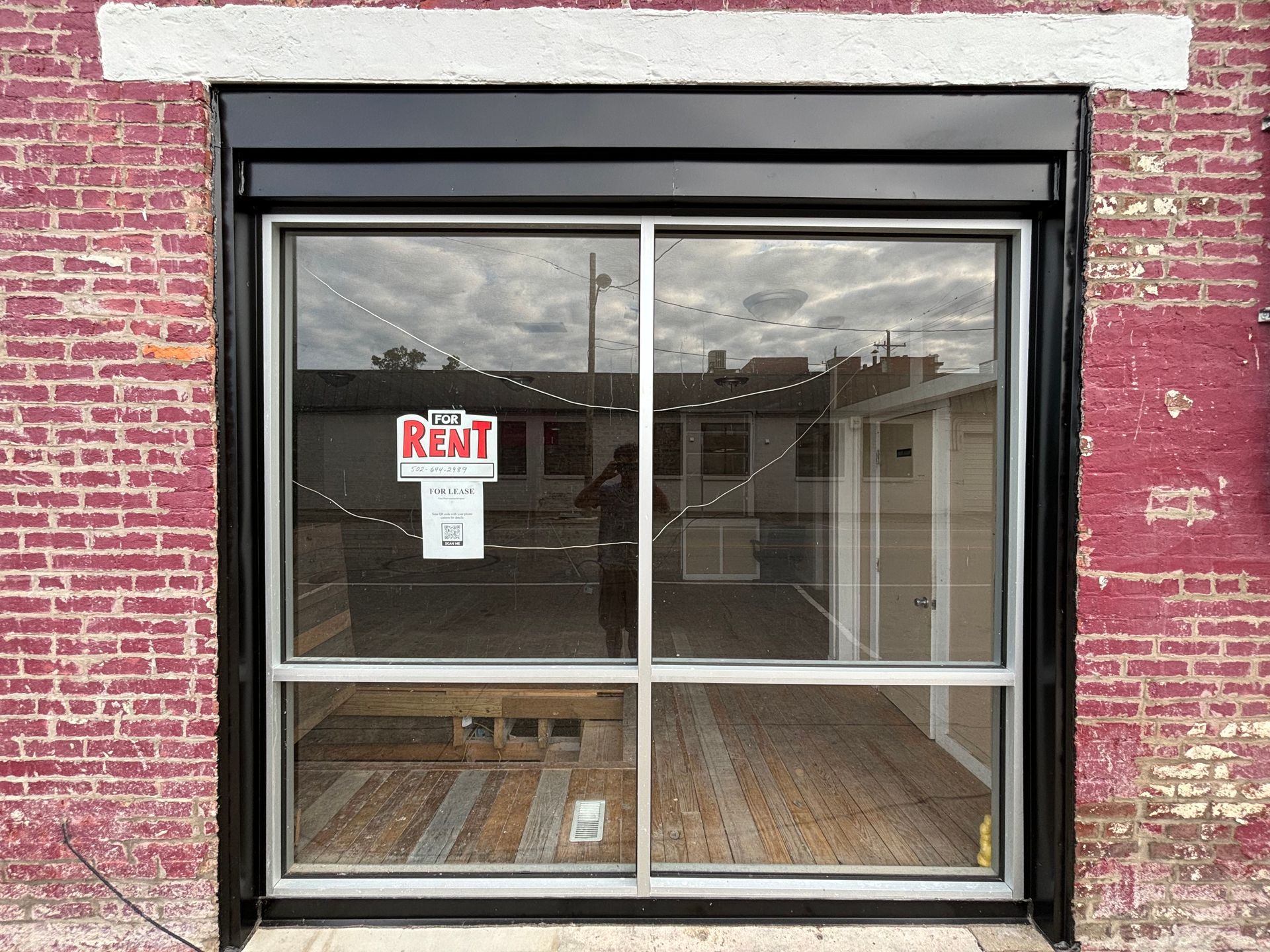 Vacant storefront with red brick facade and a large glass window displaying a “RENT” sign.