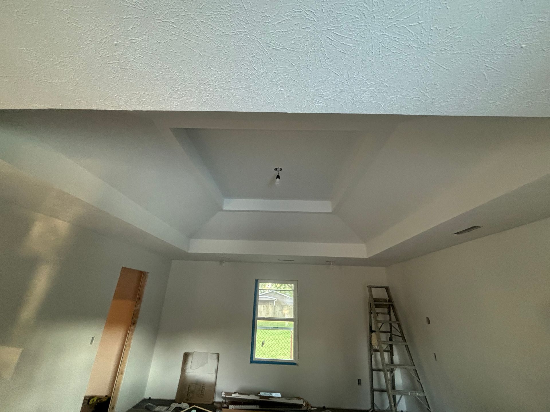 Empty room under renovation with ladder, window, and tray ceiling.