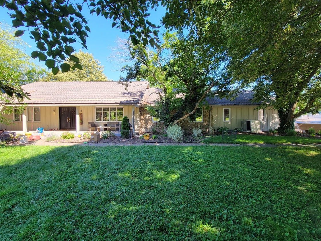 Single-story beige house with a large green lawn and leafy trees in front