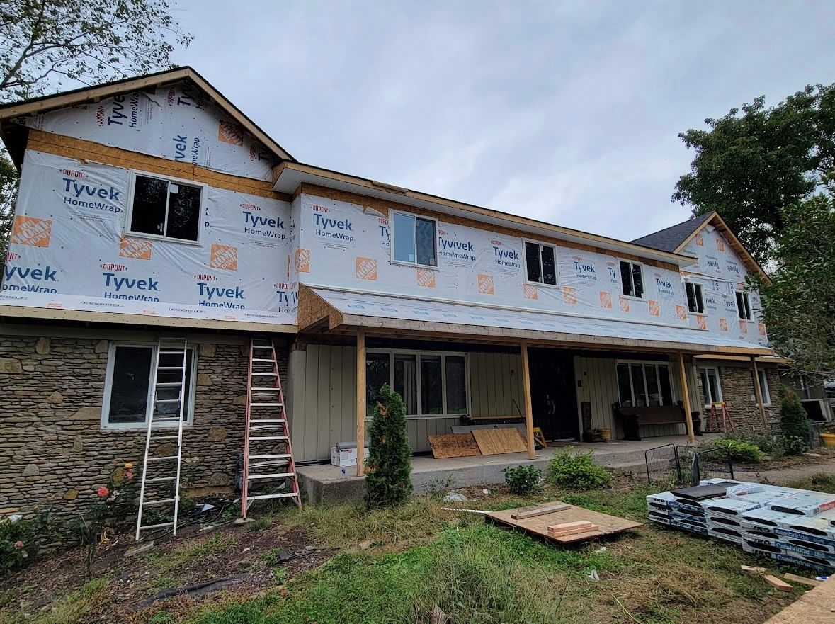 Partially renovated two-story house wrapped in Tyvek with ladders and construction materials in front