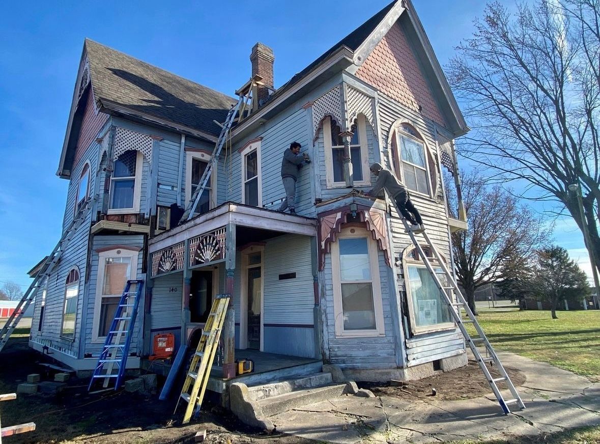 Dilapidated blue Victorian house with damaged porch and ladders in front, seen on a sunny day