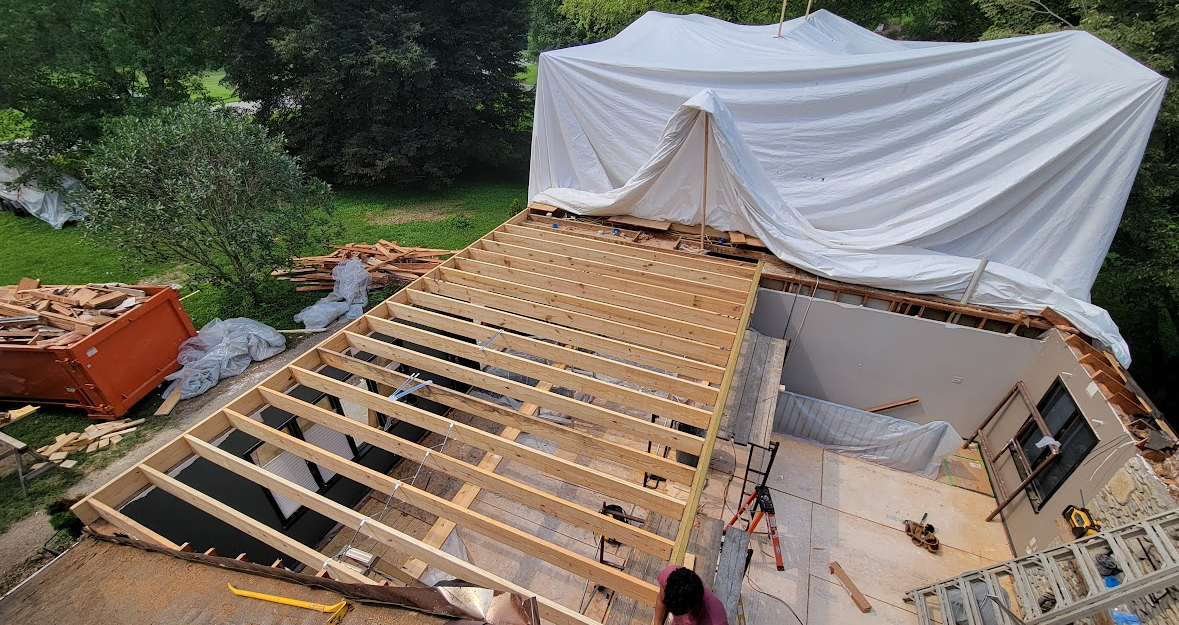 Roof construction with exposed wooden rafters beside a white tarp-covered structure outdoors