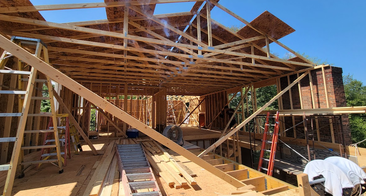 Partially framed house under construction with roof trusses, lumber, ladders, and a worker at the right edge