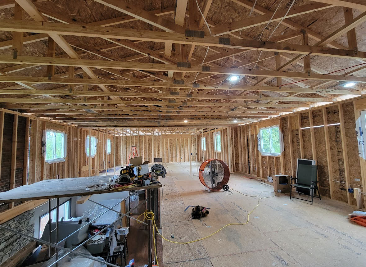Unfinished framed room with exposed wood trusses, windows, construction tools, and a fan on the floor