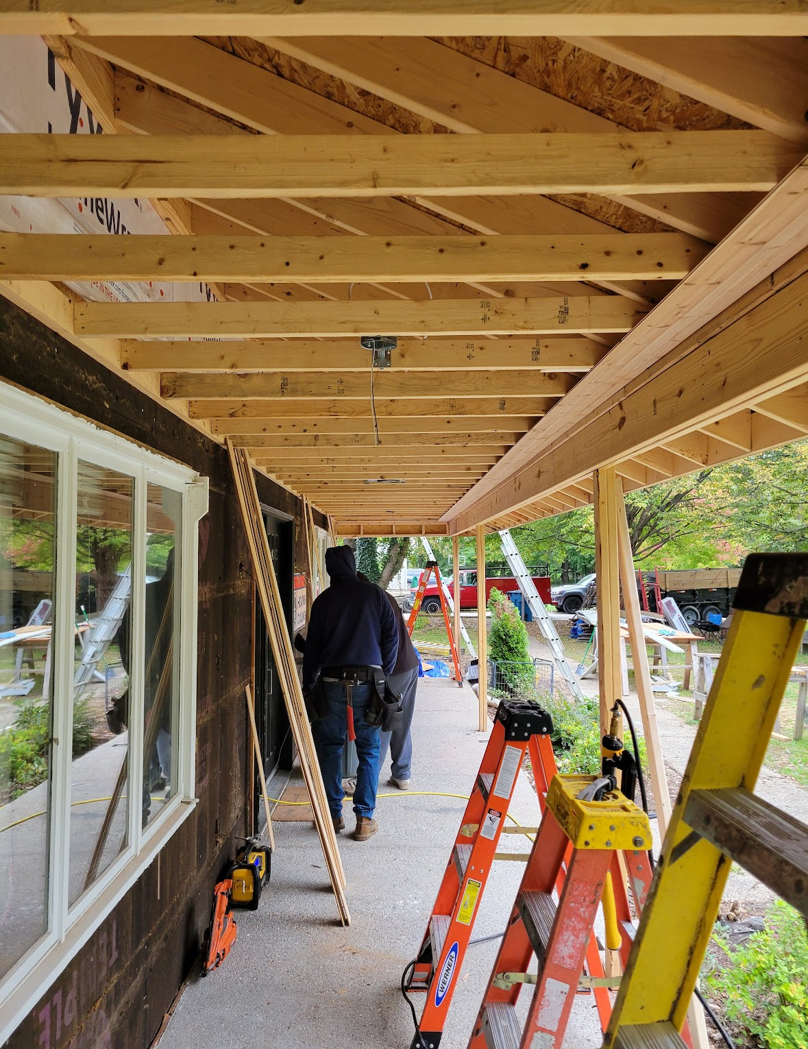 Covered porch under construction with exposed joists, ladders, and a worker in the background.