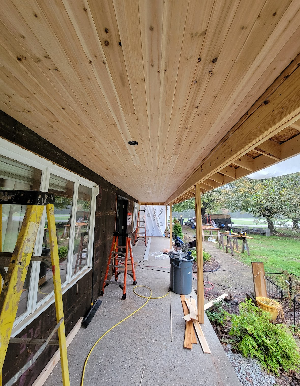 Covered porch under construction with wooden ceiling, ladders, tools, and yard visible beside the house