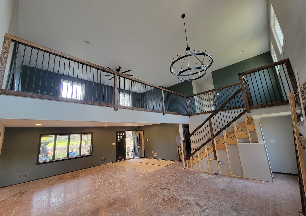 Empty two-story loft interior with staircase, balcony railing, and chandelier, under renovation
