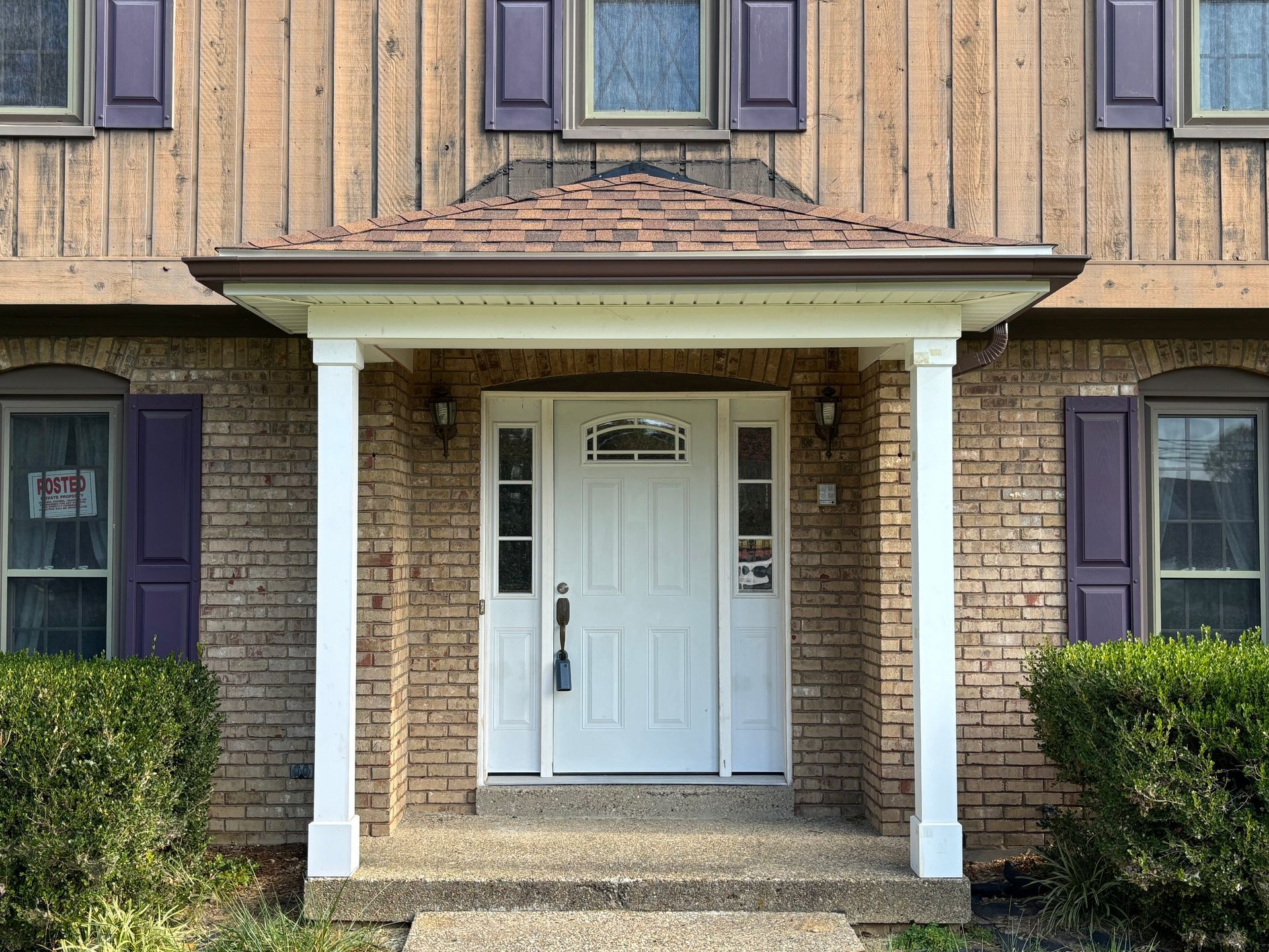 Front entrance of a brick house with white double doors, porch columns, and trimmed hedges