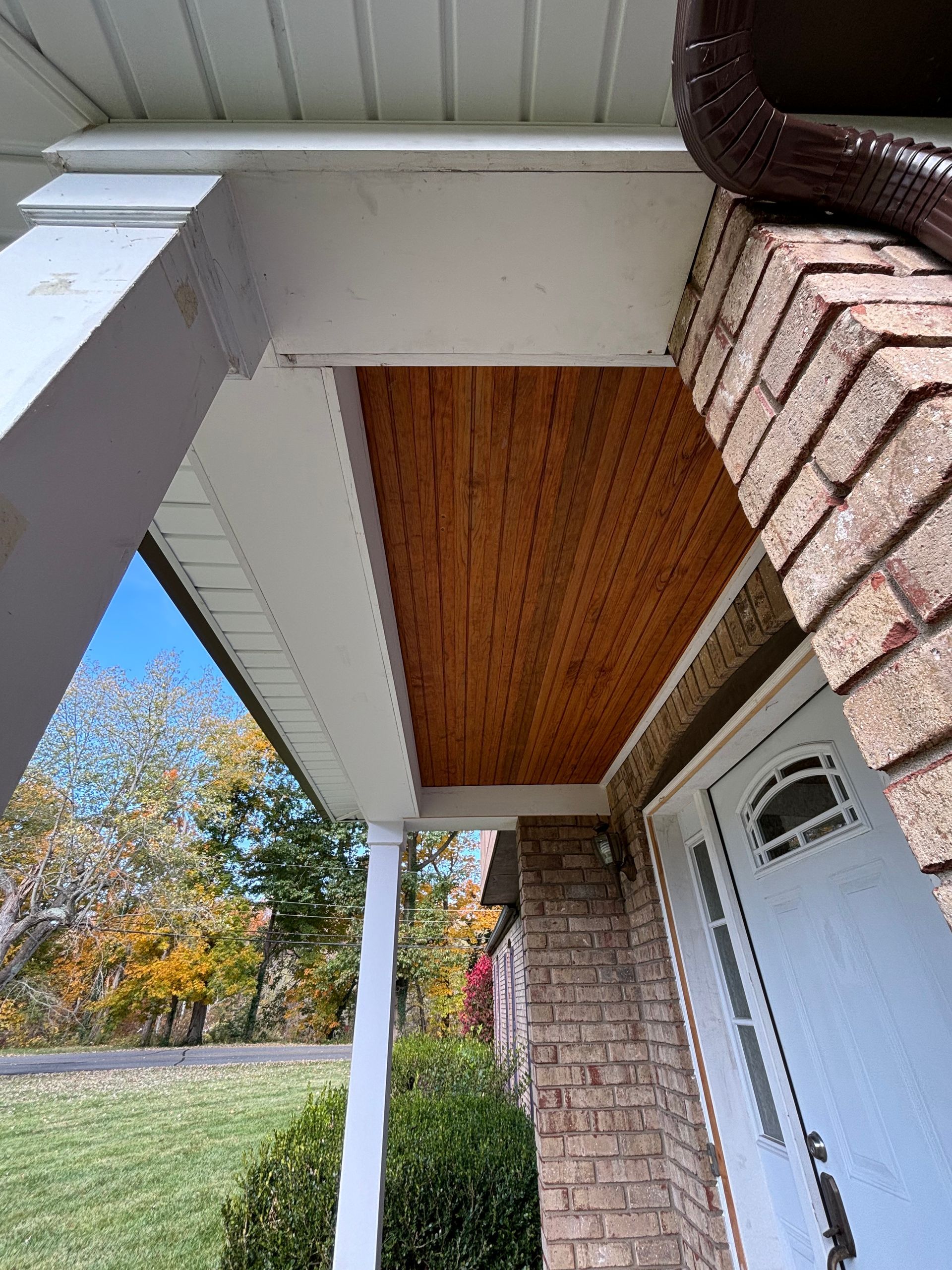 Front porch with white trim, brick siding, wooden ceiling, and a white front door beside a grassy yard.