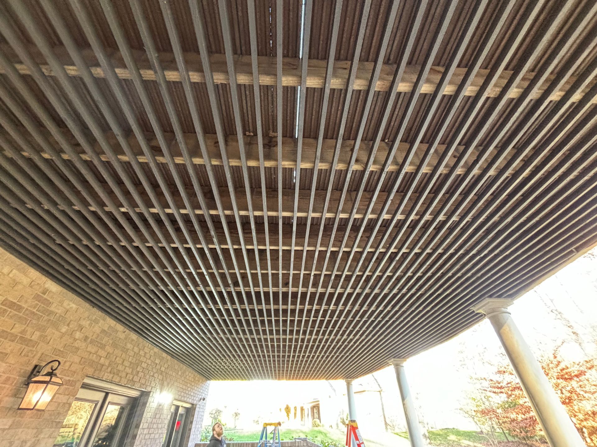 Covered patio with a slatted wooden ceiling, brick wall, and bright outdoor view beyond white columns