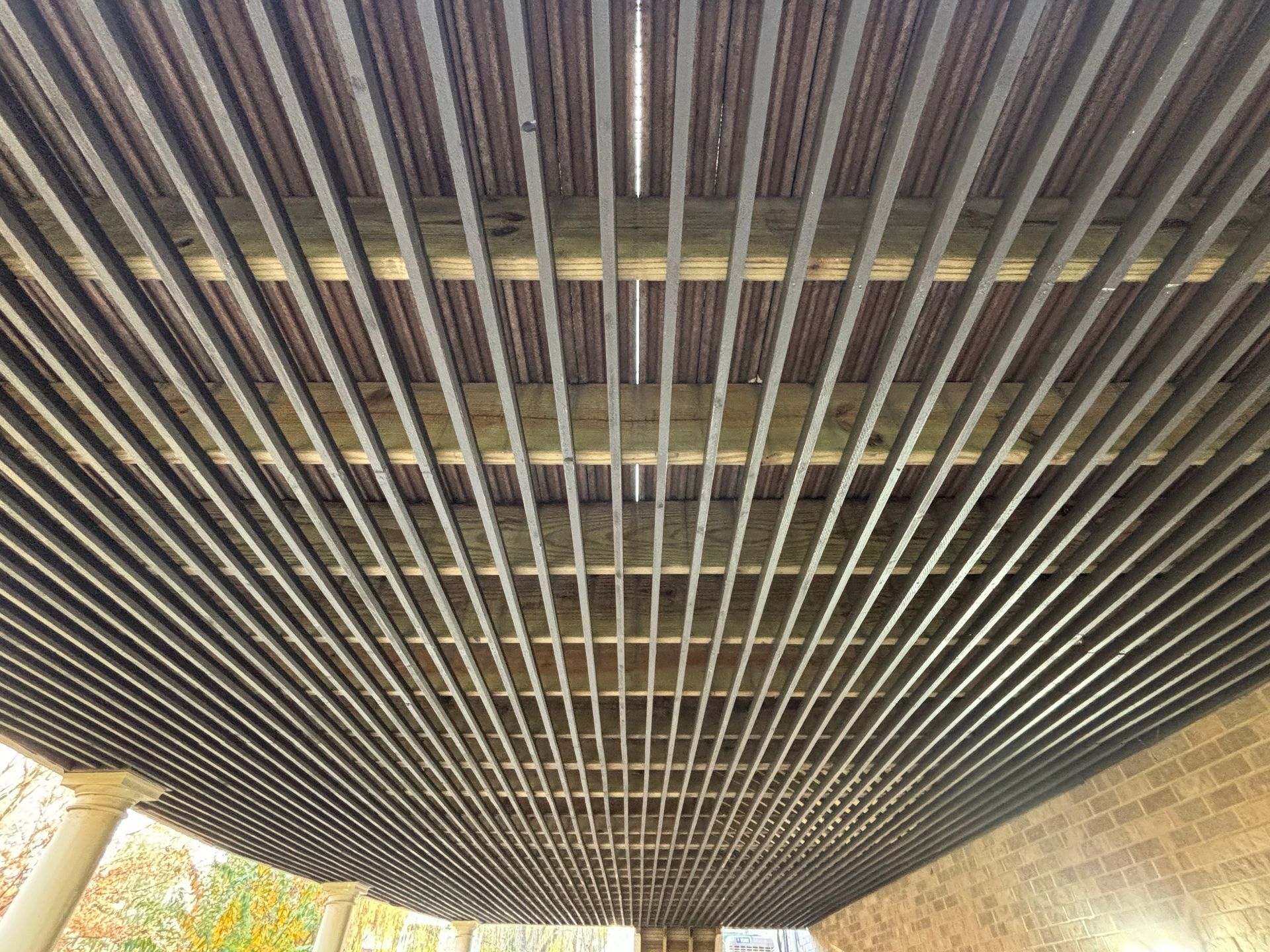 Underside of a slatted wooden canopy with sunlight filtering through and trees visible at the edges