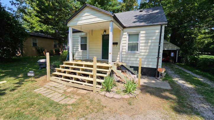 Small white house with a front porch and wooden ramp, surrounded by trees and a grassy yard.