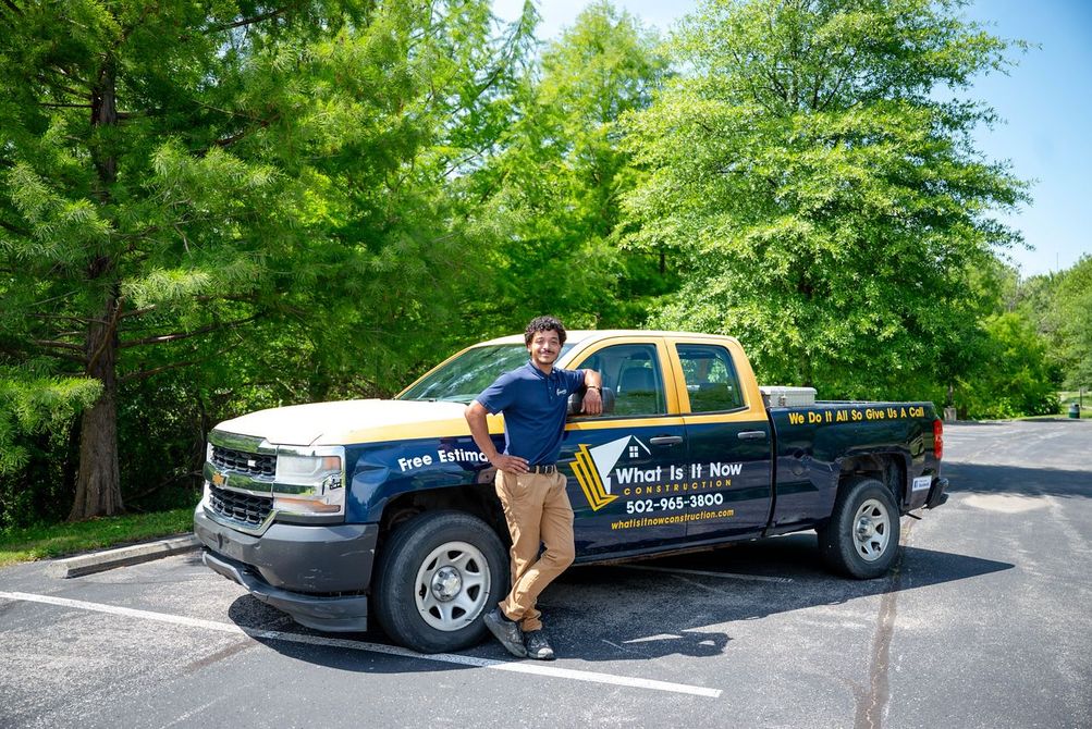 Man standing beside a black pickup truck in a sunny parking lot with green trees behind it