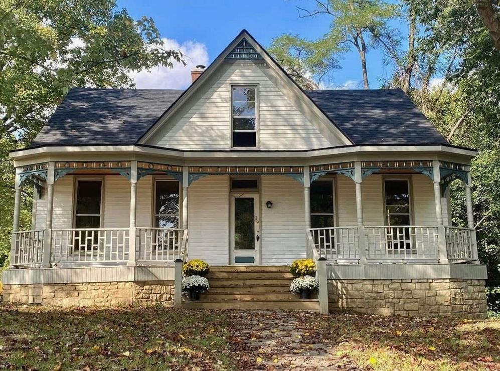 Front view of a white farmhouse with a porch, black shutters, and a steep gabled roof amid autumn trees