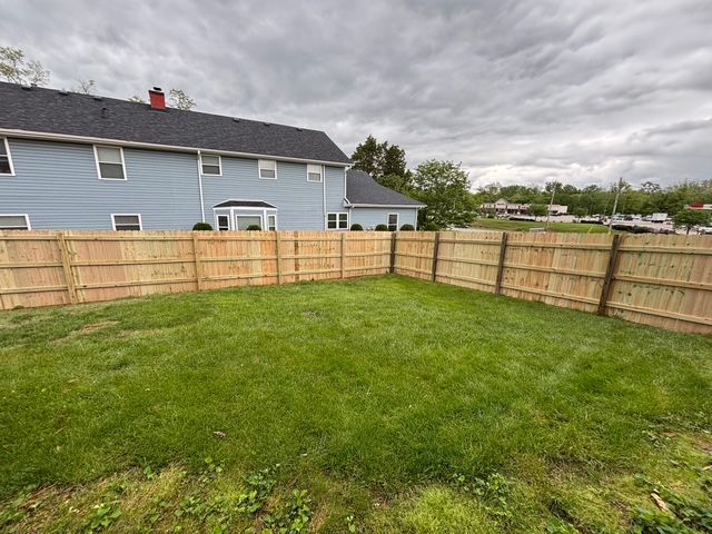 Grassy backyard with a wooden fence and a blue house under cloudy skies.