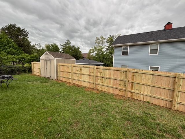 Backyard with wooden fence, storage shed, and blue house under cloudy sky