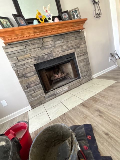 Stone fireplace with wooden mantel in a tiled living room, with a chair and bucket in the foreground