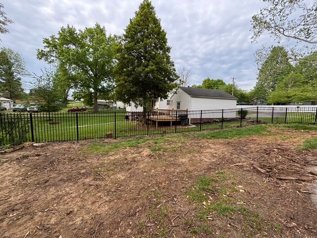 Fenced yard with bare dirt, trees, and a white barn under a cloudy sky