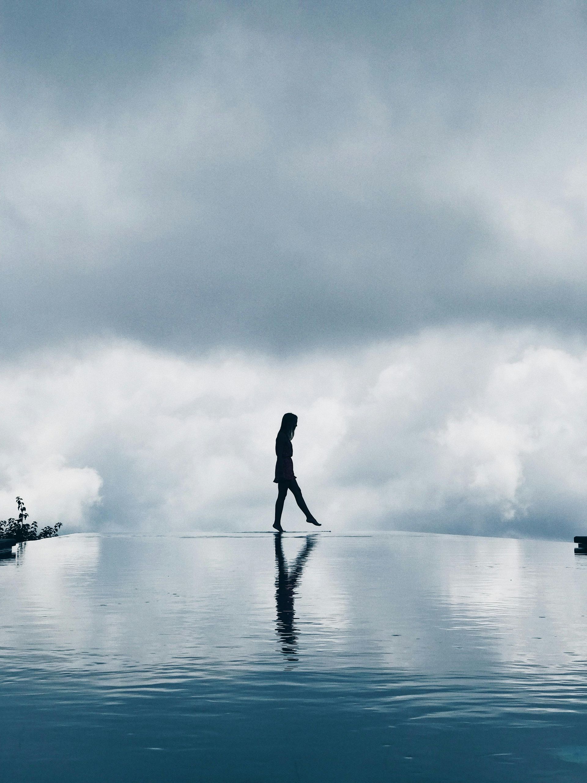 Person walking on water, reflecting in calm surface, cloudy sky backdrop.