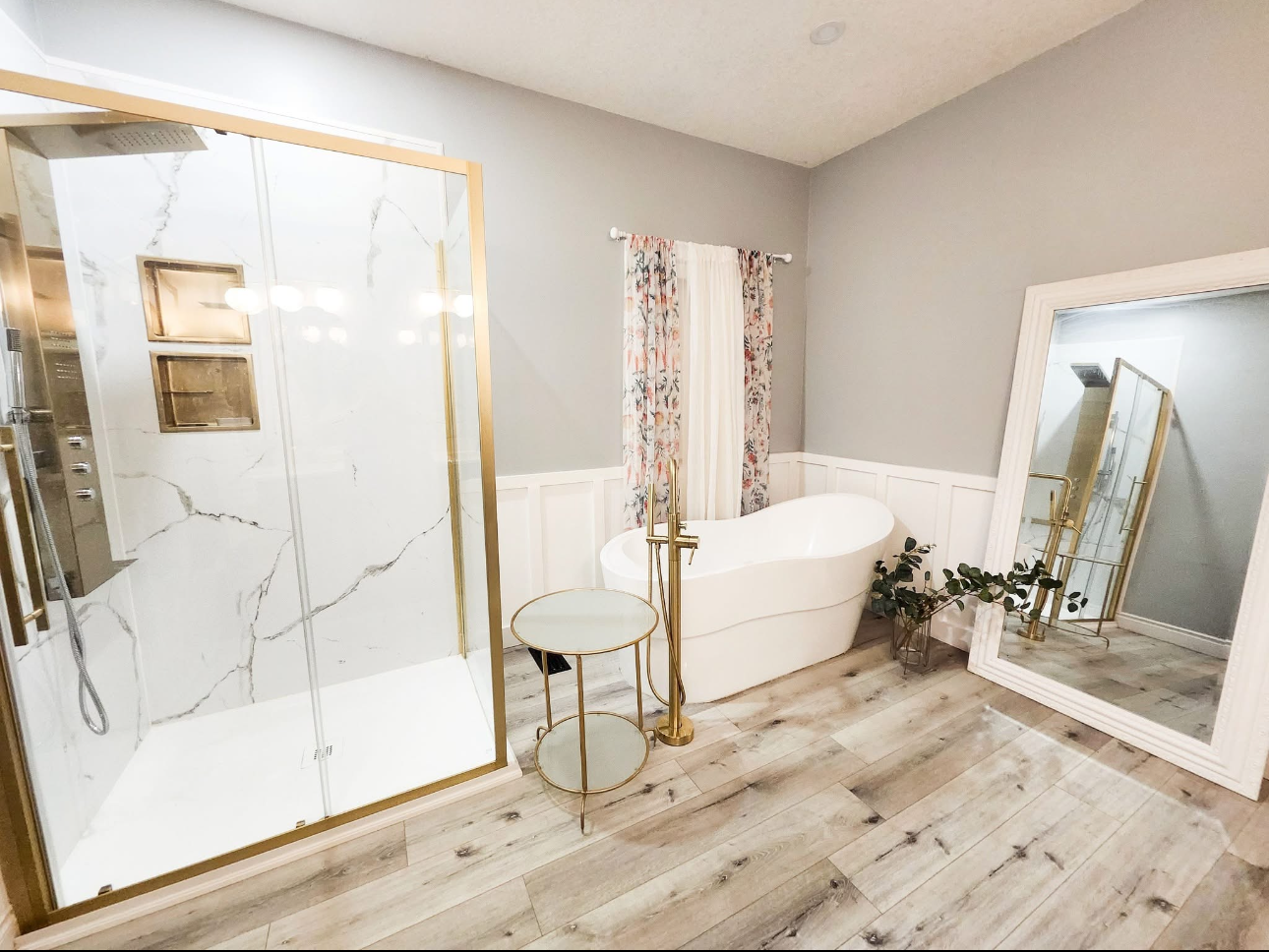 Luxury bathroom with gold accents, marble shower, freestanding tub, large mirror, and light wood flooring.