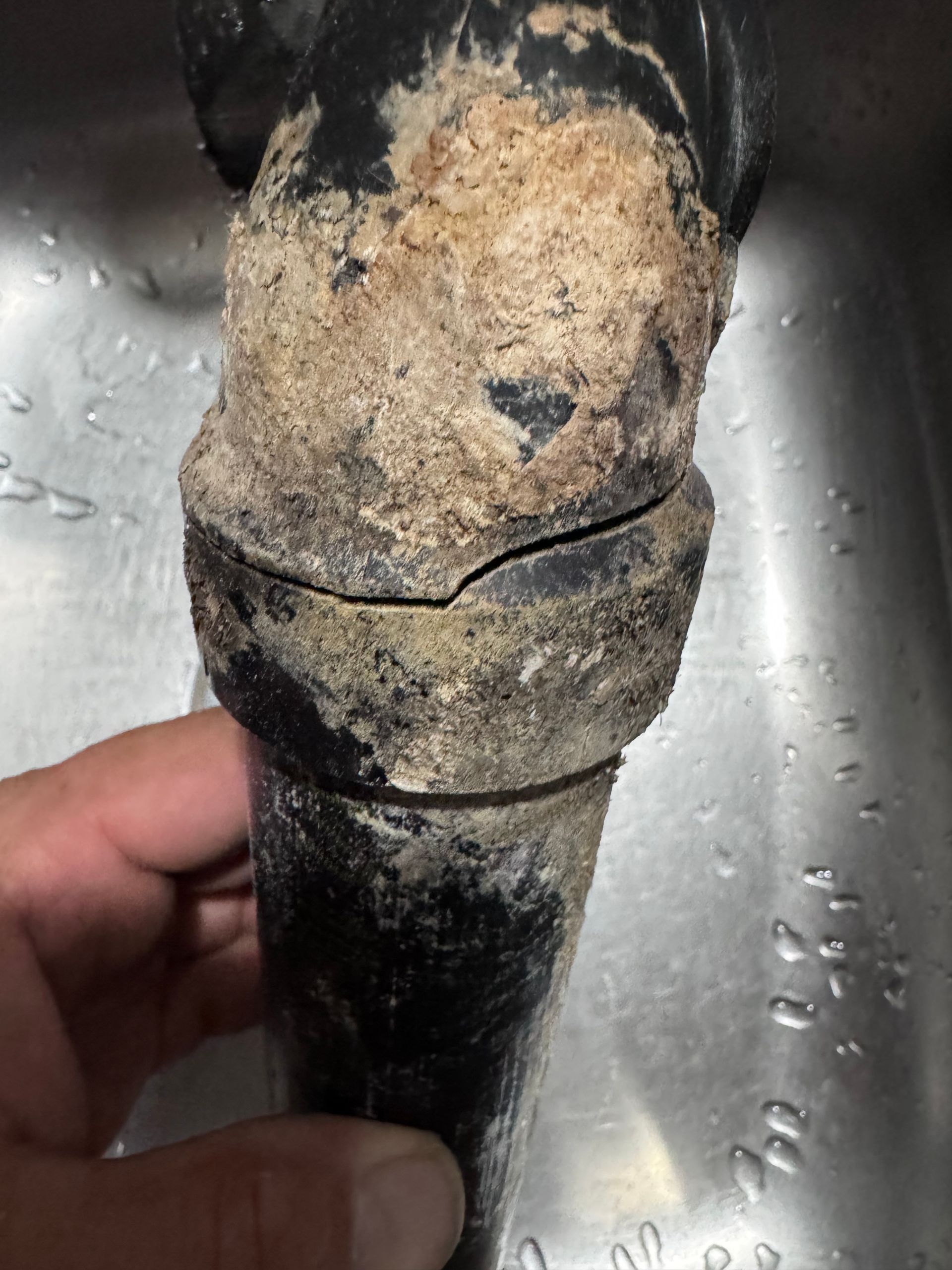 Cracked black plumbing pipe with significant mineral buildup. Hand holding the pipe against a stainless steel sink.