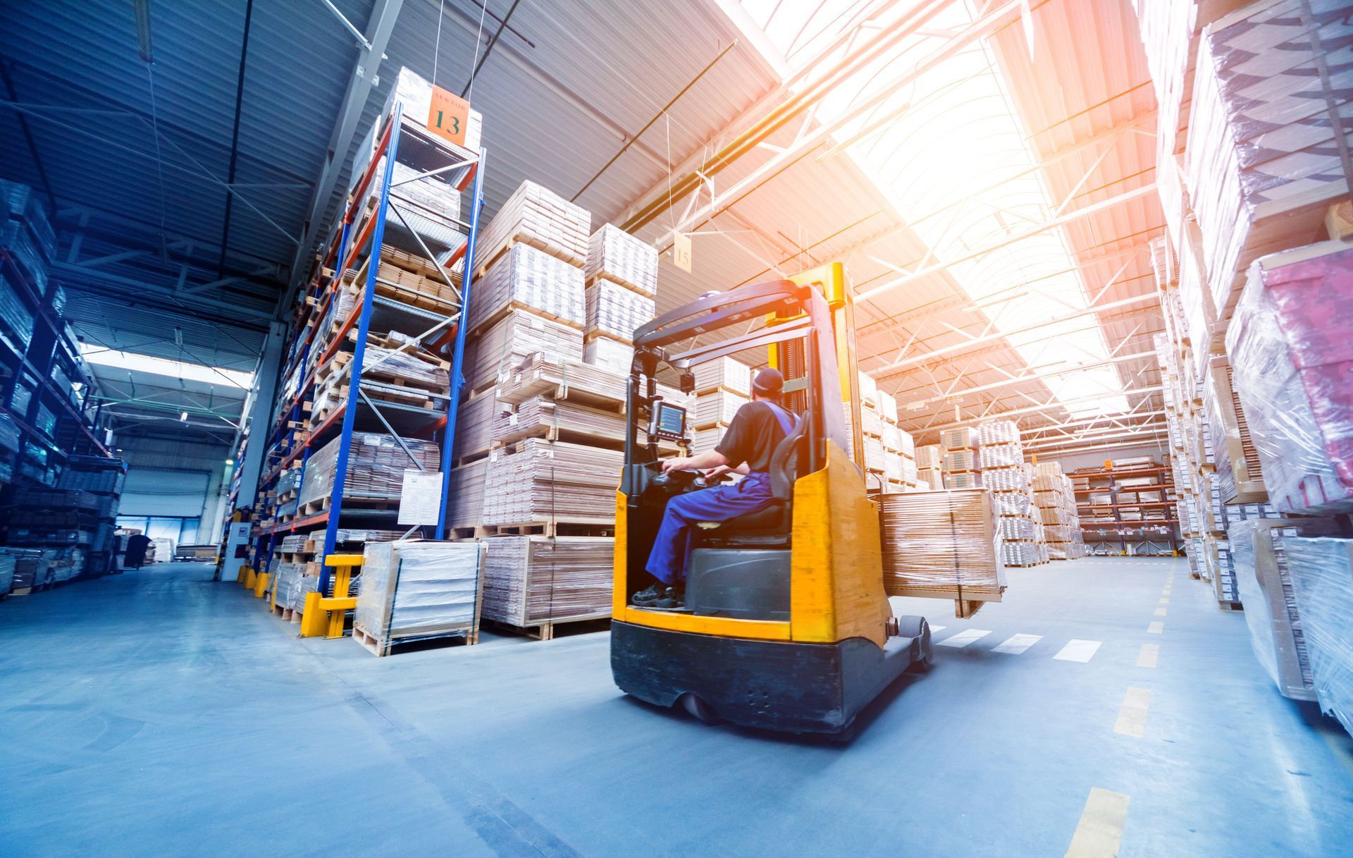 Forklift driving through a warehouse aisle with stacked pallets and bright overhead light.