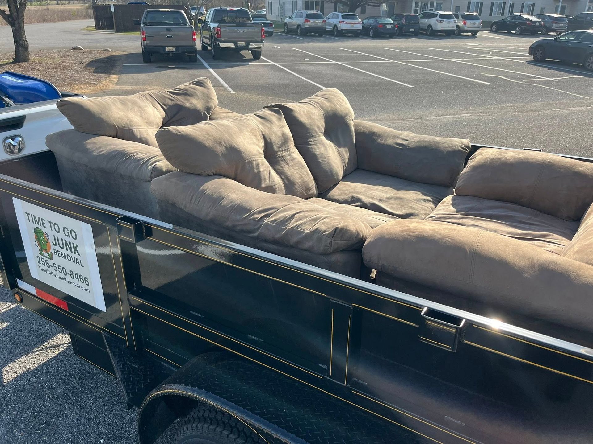 A tan sofa sits in a black utility trailer parked in a lot, with a branded sign on the side.