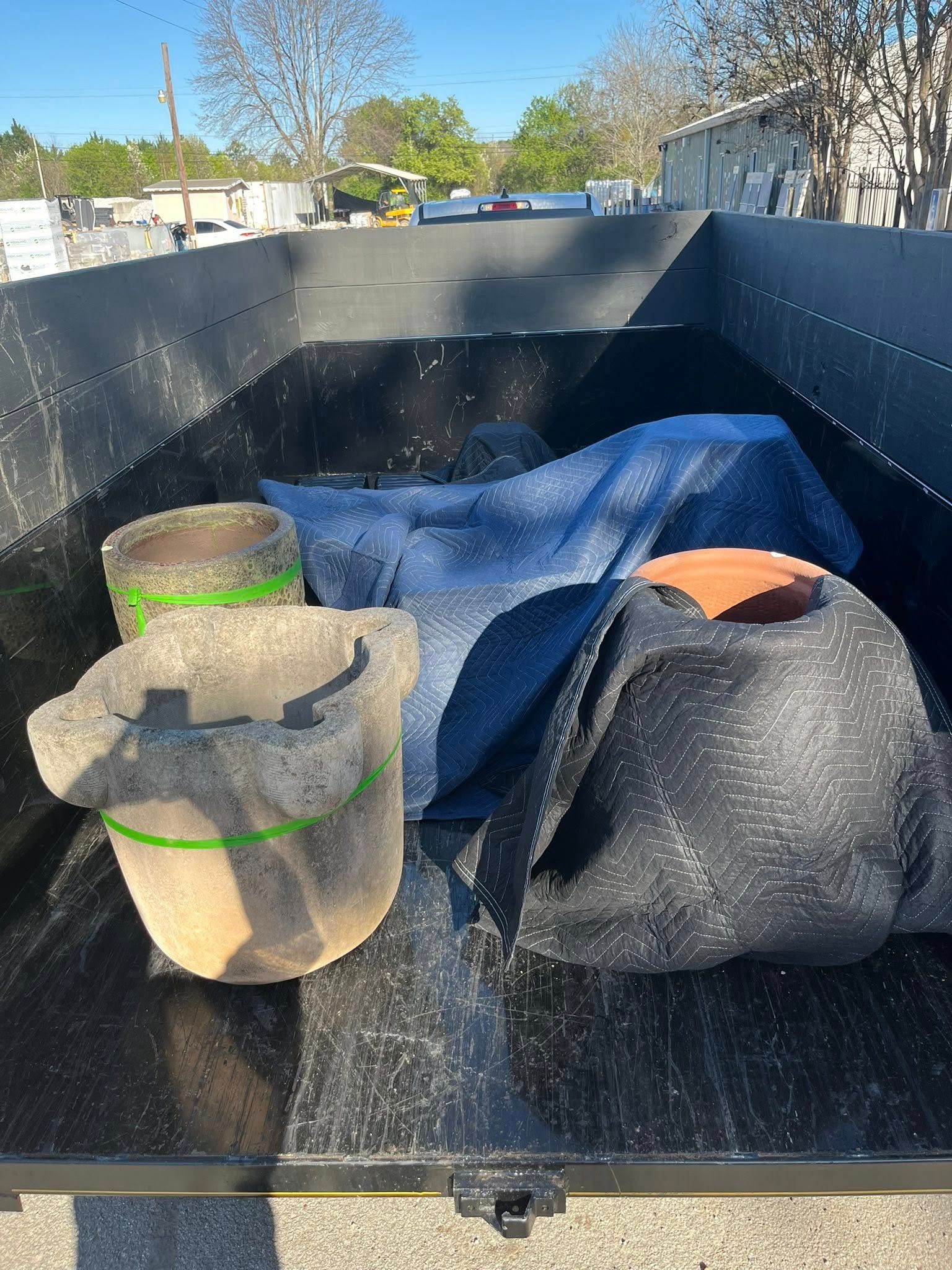 Two large, textured outdoor pots and a blue fabric tarp sitting in the bed of a black trailer on a sunny day.