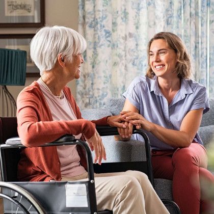Two women smiling and holding hands in a care setting, one seated in a wheelchair