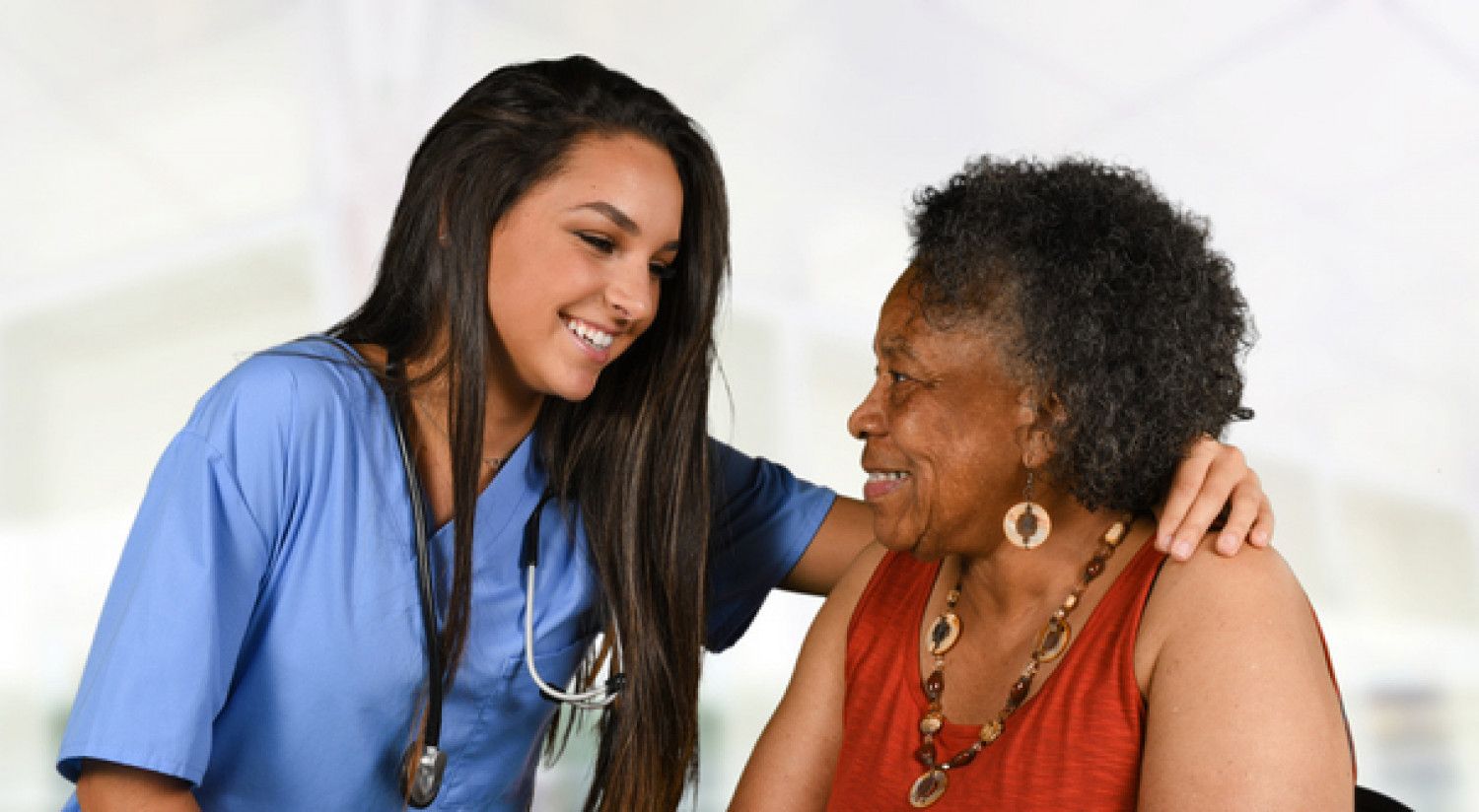 Healthcare worker in blue scrubs smiles and talks with a seated patient in a bright clinic room